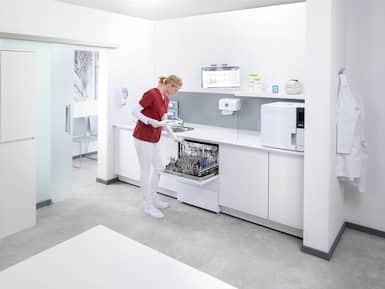 Helsearbeider i røde operasjonsklær utensiler medisinske/tannlegeinstrumenter av metall i en sterilgods-vaskemaskin i et rent, hvitt rom med skap og utstyr for instrumentbehandling på en klinikk.
