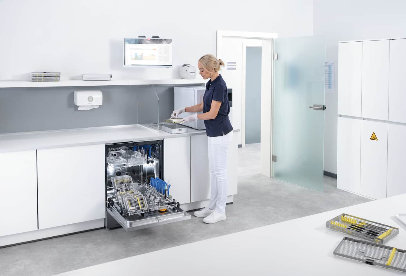 Modern sterile clinic reprocessing room; gloved staff handles instrument trays by an open washer-disinfector.