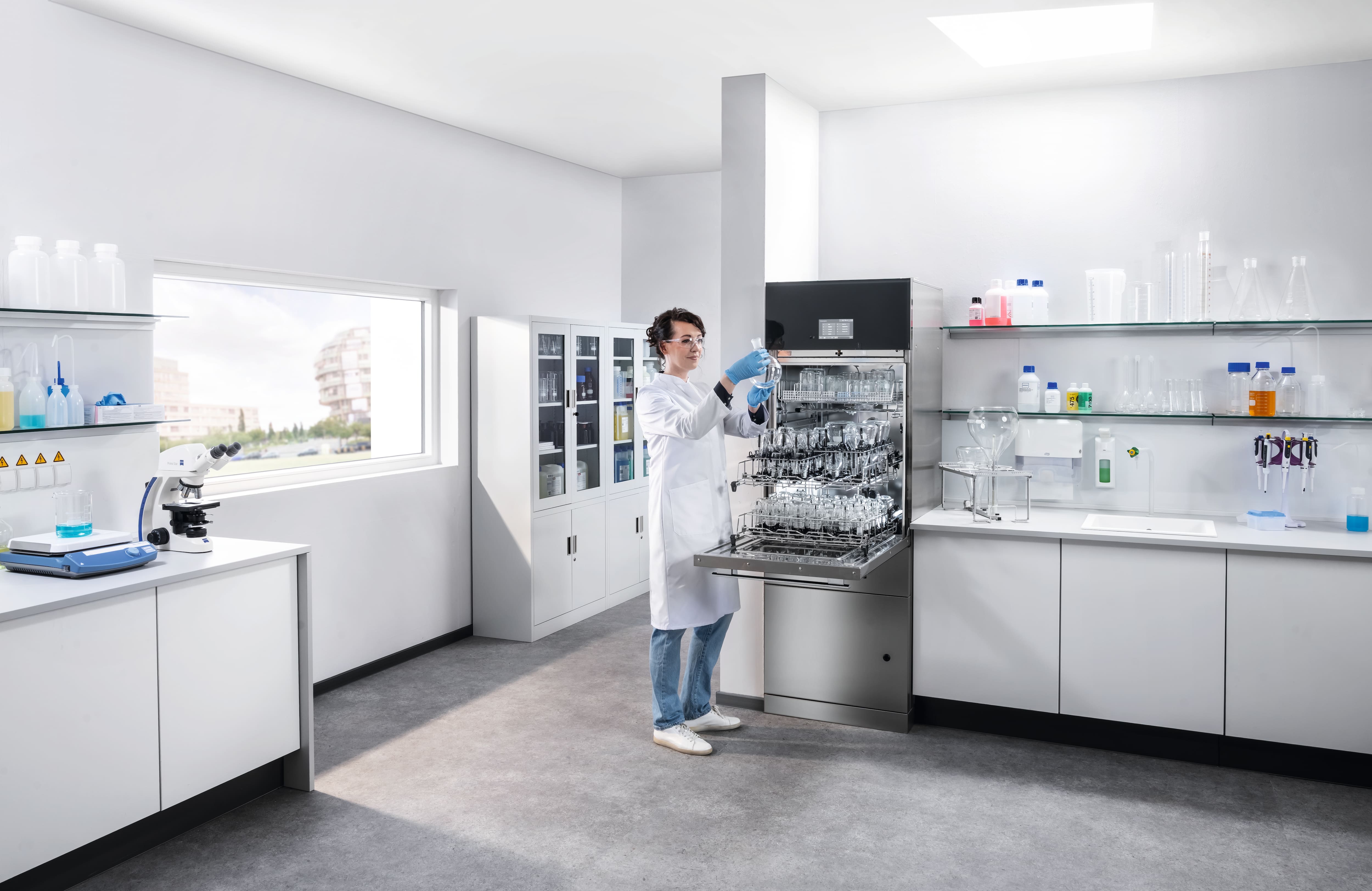 A lab technician is standing next to a SlimLine lab glasswasher, holding a clean round-bottom flask.