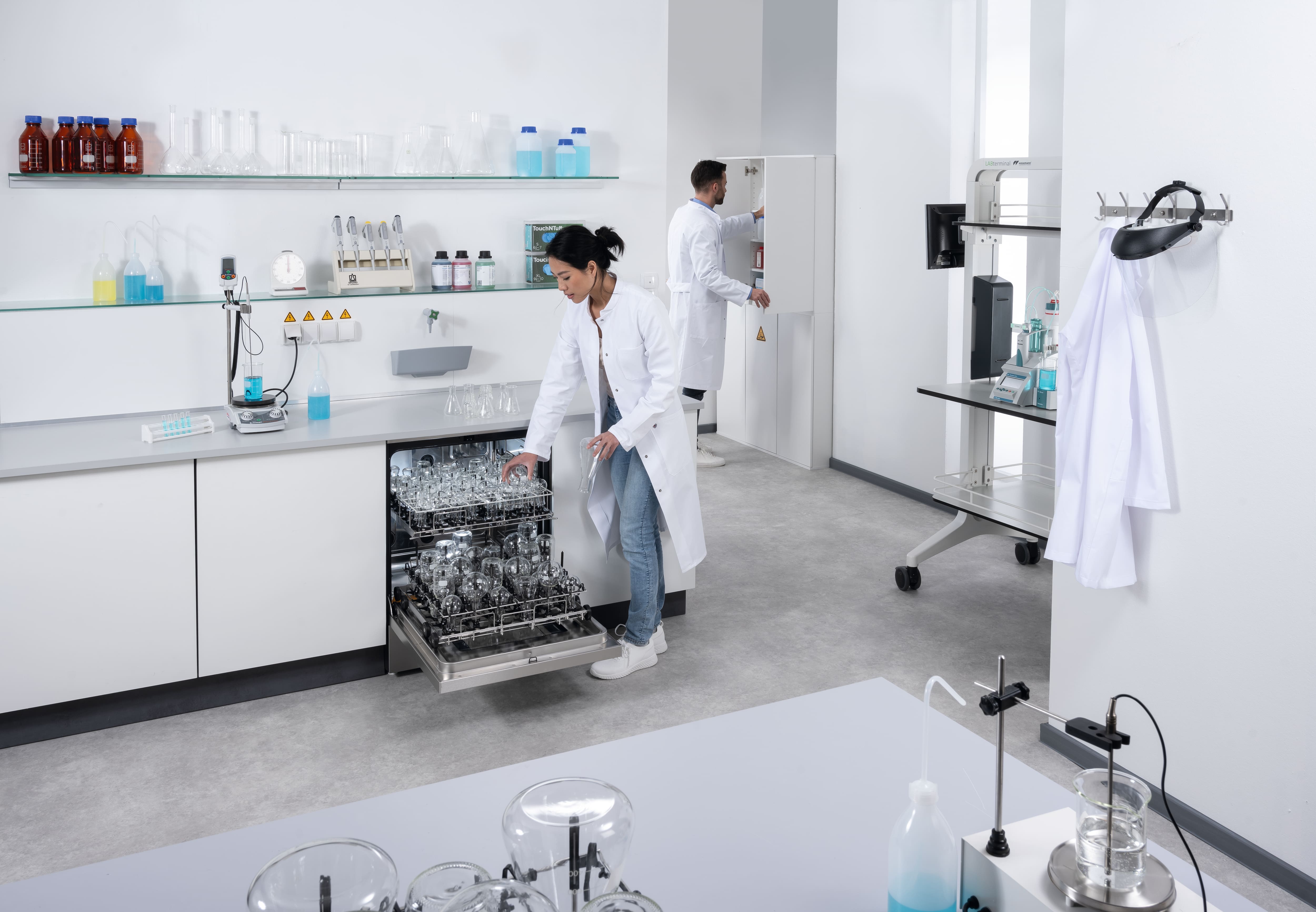Une technicienne de laboratoire, vêtue d'une blouse, place de la verrerie de laboratoire dans un lave-vaisselle de laboratoire encastré sous le plan de travail, dont la porte est ouverte.